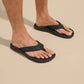 Men's Olukai Ohana black flip flop sandals worn on male feet against beige background