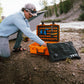 Person kneeling outdoors opening an orange YETI Loadout GoBox with protective KOR inserts near a river