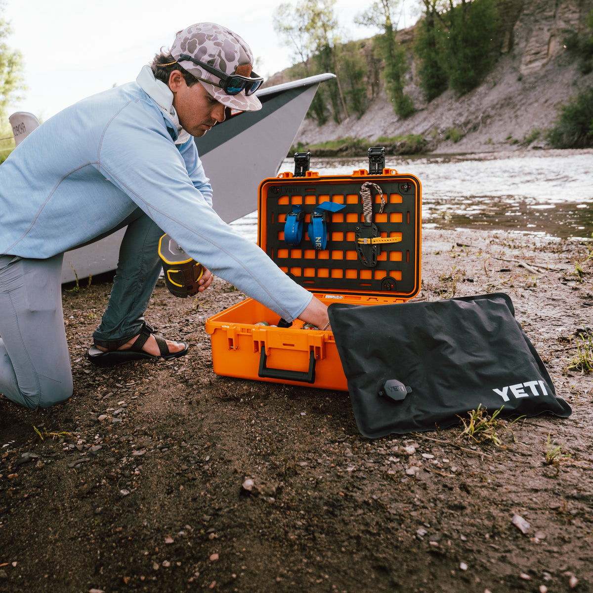 Person kneeling outdoors opening an orange YETI Loadout GoBox with protective KOR inserts near a river