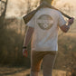 Man carrying shotgun wearing Local Boy Outfitters Graphic Vintage Sphere Sand Tee outdoors at golden hour
