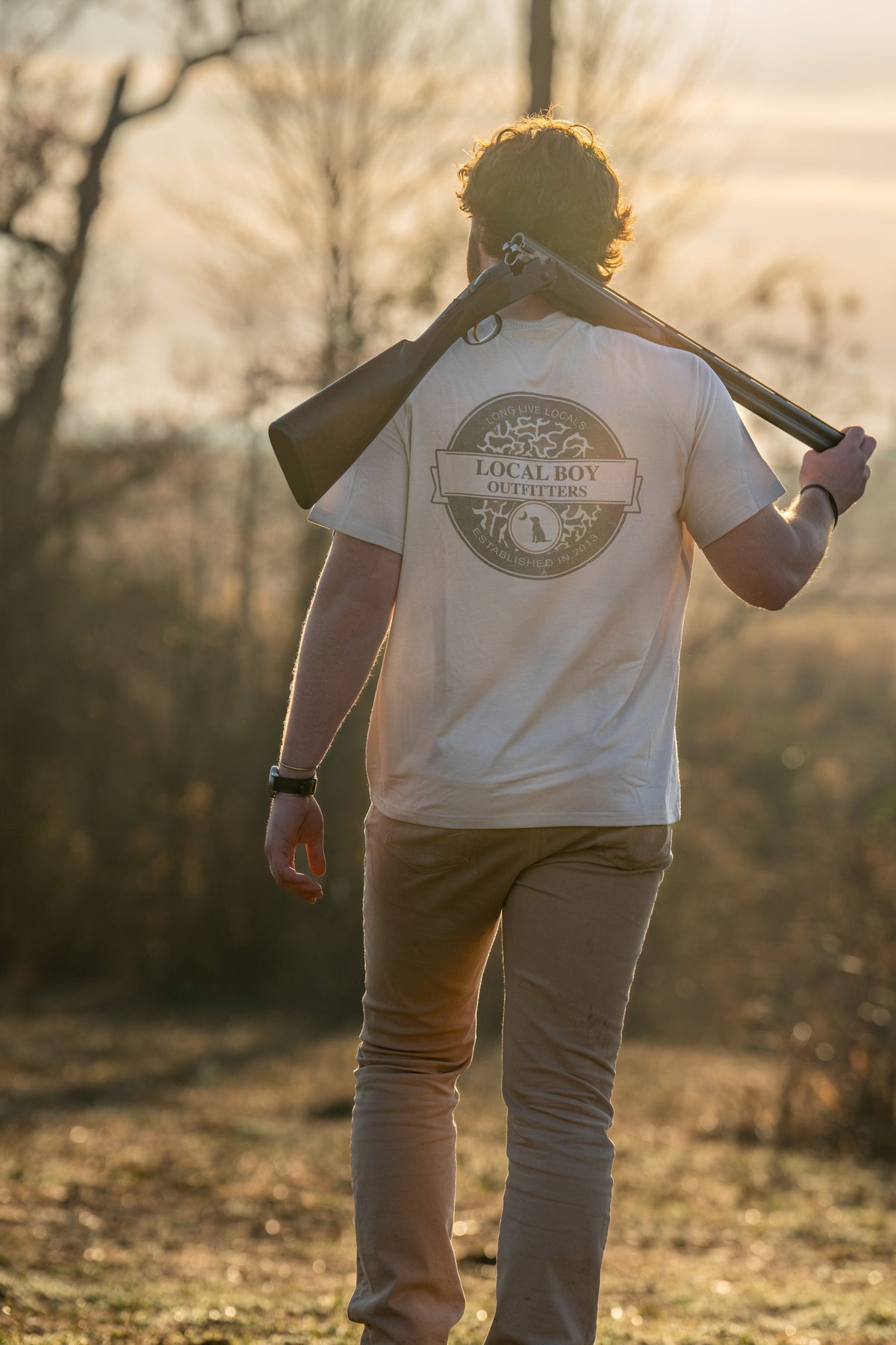 Man carrying shotgun wearing Local Boy Outfitters Graphic Vintage Sphere Sand Tee outdoors at golden hour