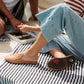 OluKai Women's Mi'i Mule in Kukui Brown woven leather on striped beach towel