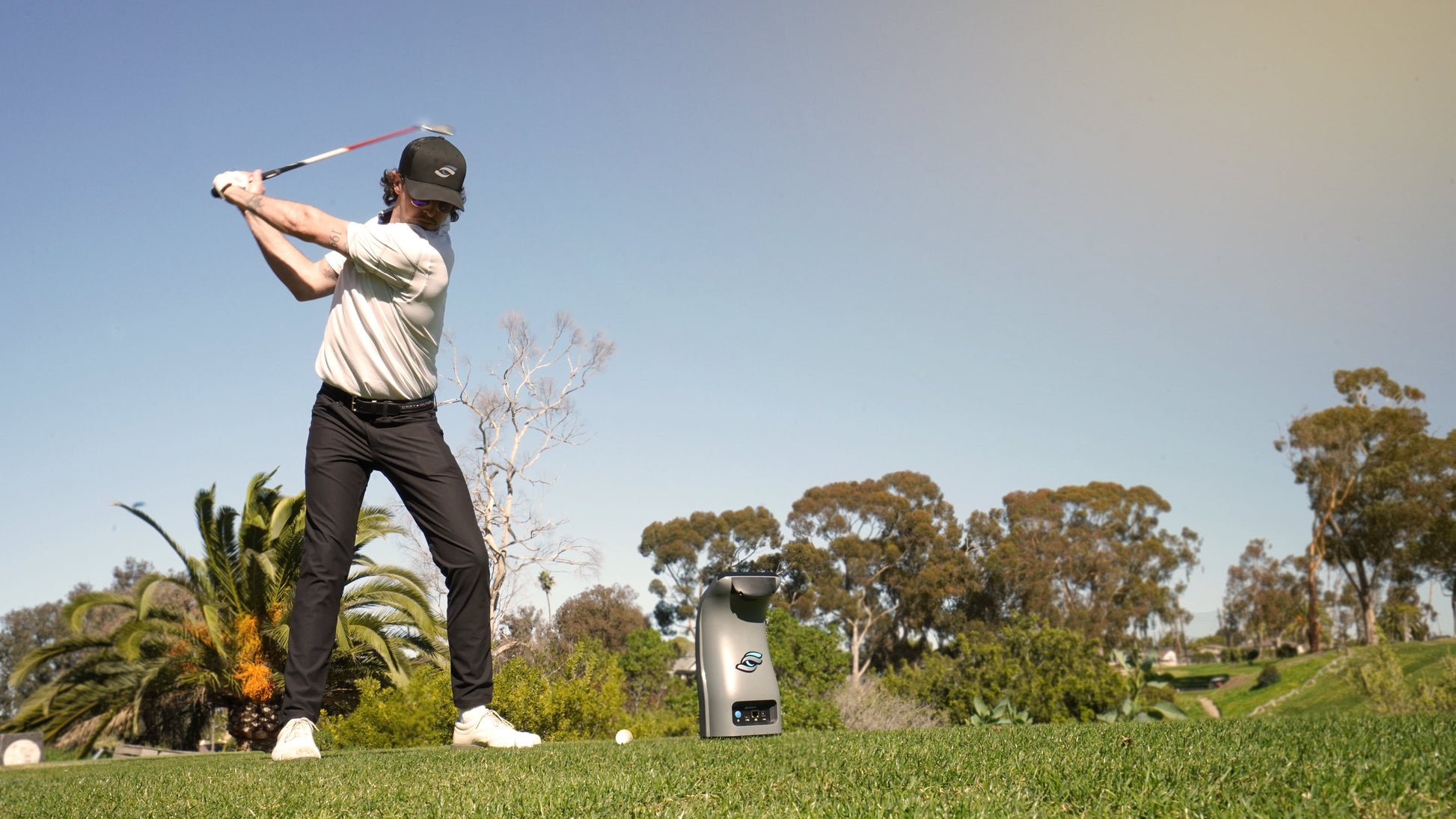 Golfer swinging club on course with Foresight QC3 launch monitor device placed nearby on grass