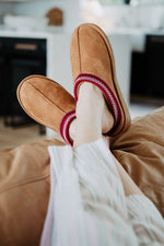 Katydid Chestnut cozy slippers with red embroidered trim worn on feet resting on sofa
